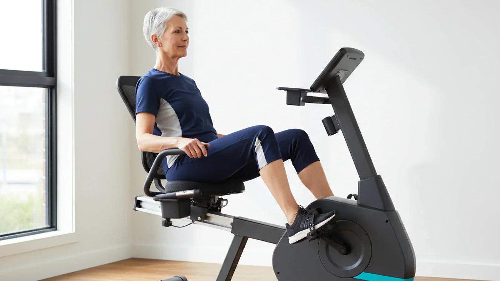 Mature Australian woman seated on a charcoal-grey recumbent exercise bike in a sunlit home gym, showing the reclined mesh backrest, step-through frame, slide rail and console with tablet shelf